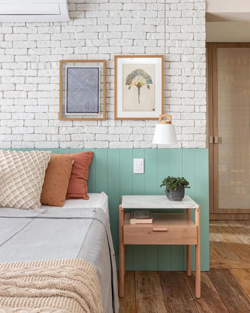 Parede de tijolinhos brancos e com painel de madeira pintado de verde água na metade, também há uma cama com almofadas, mesa de cabeceira com vaso de planta e quadros na parede