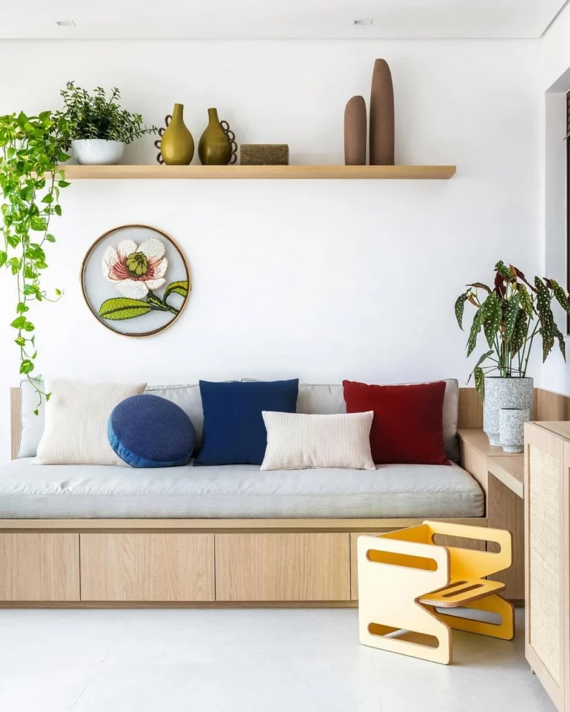 Parede branca decorada com prateleira e itens decorativos, além de um quadro bordado; também há um sofá moderno com almofadas coloridas em tons de azul, branco e bordô.