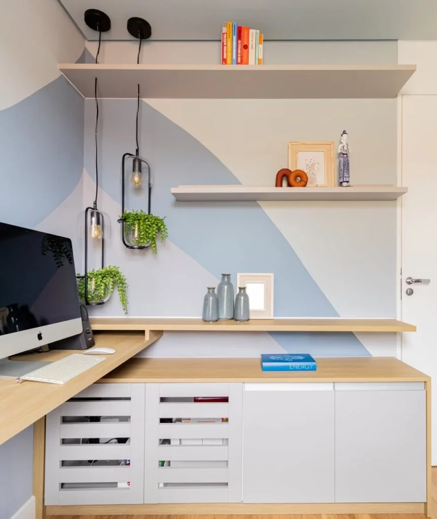 Espaço de home office em casa com parede pintada em tons de azul, plantas penduradas, prateleiras com acessórios decorativos e computador na mesa.