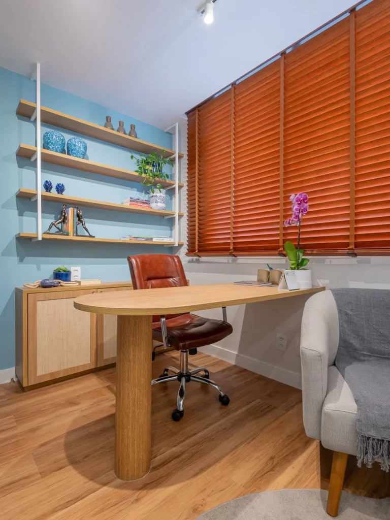 Escritório com parede azul, persianas terracota, prateleiras com objetos decorativos, mesa com vaso de flor e cadeira de couro marrom.