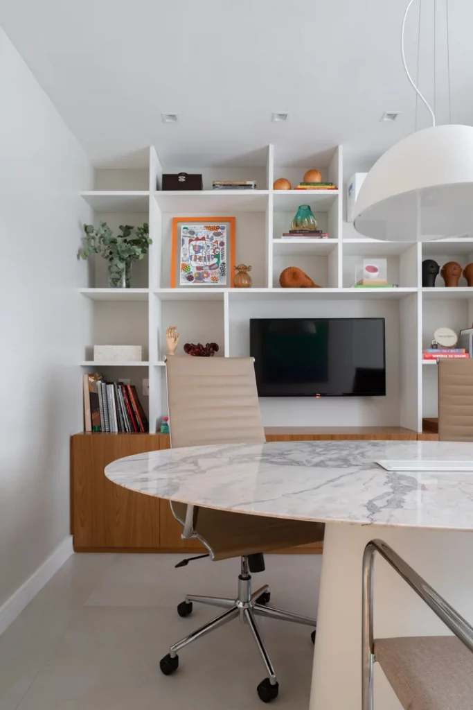 Escritório com prateleira e nichos vazados brancos, com quadros decorativos, planta, aparelho de transmissão, livros, cadeira bege e mesa de mármore.