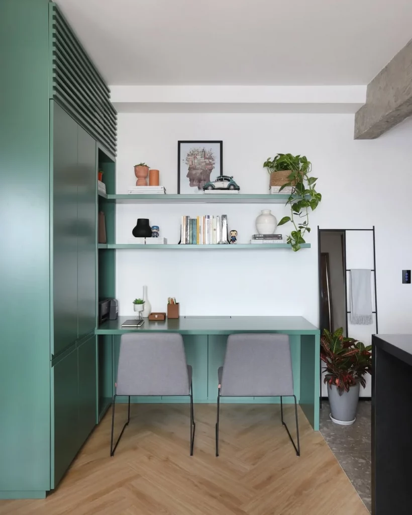 Home office compartilhado com duas cadeiras cinzas, prateleiras com plantas, acessórios decorativos e quadro, mesa e estante lateral verde escuro.