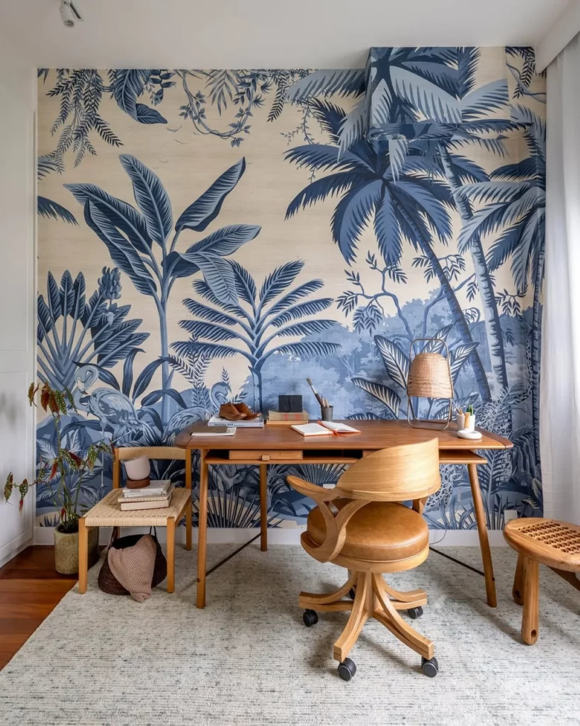 Home office com papel de parede maximalista azul e tropical, mesa e cadeira de madeira.