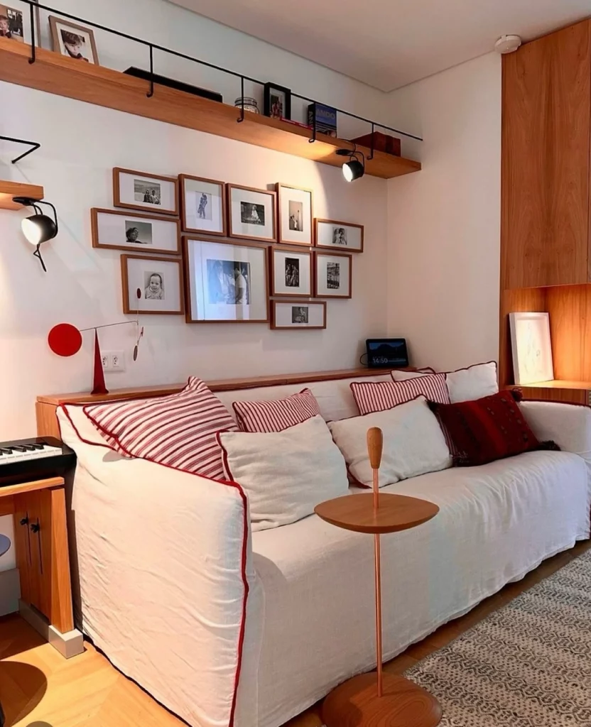 Galeria de fotos em casa com quadros dispostos em diversos tamanhos e ângulos. A sala de estar é composta de um sofá branco e almofadas vermelhas.