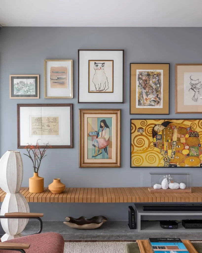 Parede cinza com mix de quadros diferentes, de cores e tamanhos diversos. Também há um aparador para enfeitar o espaço.