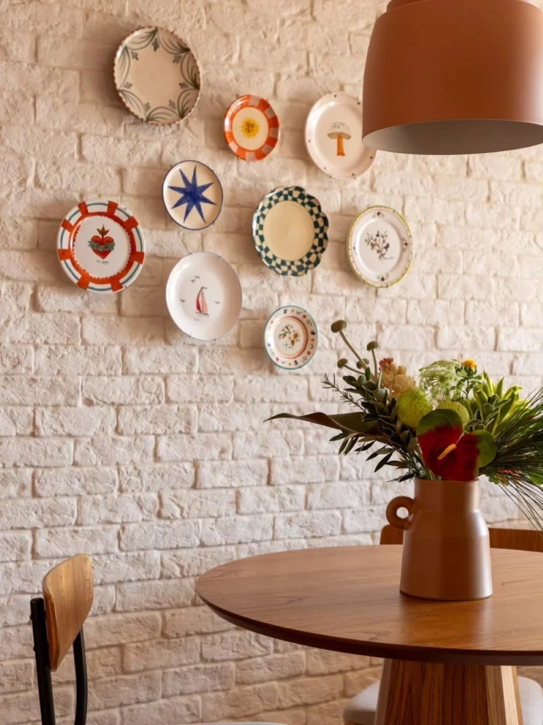 Parede de tijolinhos brancos com pratos de porcelana decorando, também há uma mesa e cadeira de madeira com um vaso de plantas