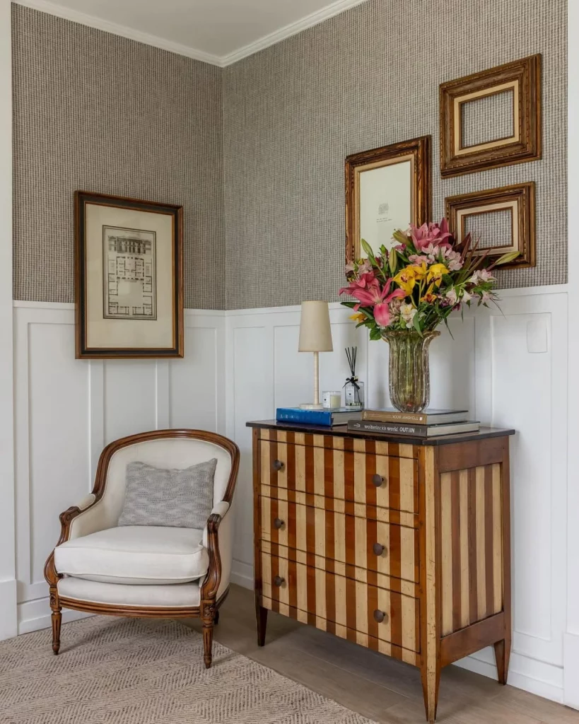 Cantinho de casa com papel de parede e boiserie, com quadros, poltrona e aparador. Estilo clássico na decoração e aconchego.