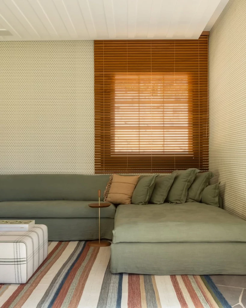 Sala com papel de parede texturizado e neutro, com sofá grande verde e moderno junto de um tapete listrado colorido
