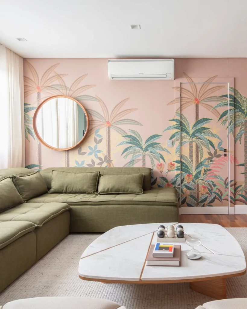 Sala com papel de parede maximalista em tons de rosa e com estampa de coqueiros; sofá verde, espelho e mesa de centro branca equilibrando o visual moderno do espaço