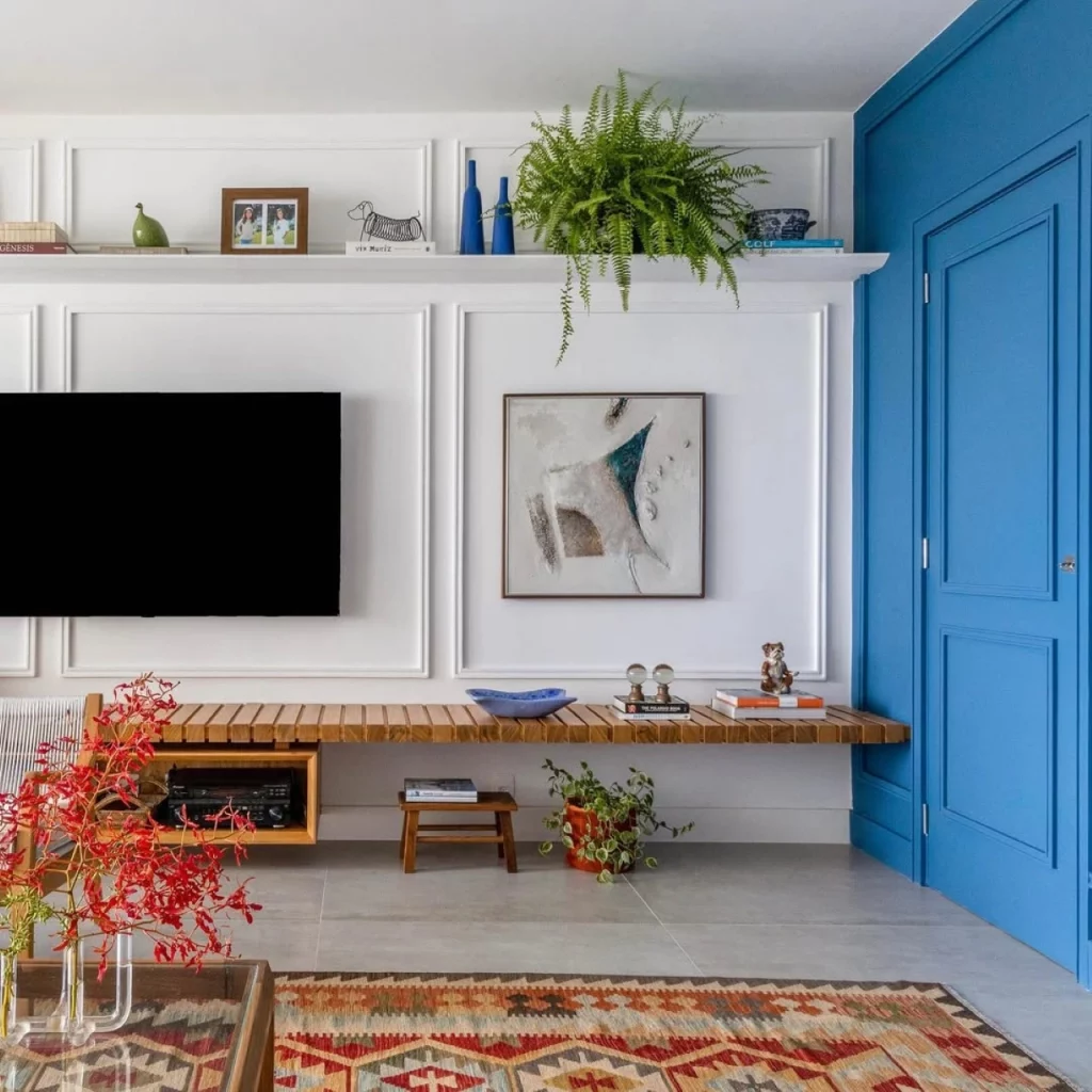 Sala de estar colorida que tem parede de boiserie, elementos decorativos como plantas, vasos e prateleiras dão alegria ao ambiente