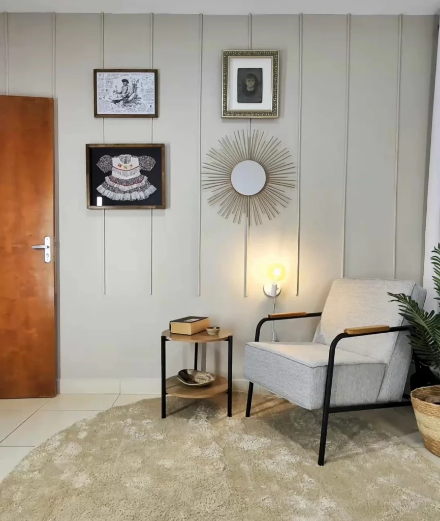 Parede decorada com canaletas no estilo clássico, com quadros complementando a decoração e poltrona branca com mesinha ao lado