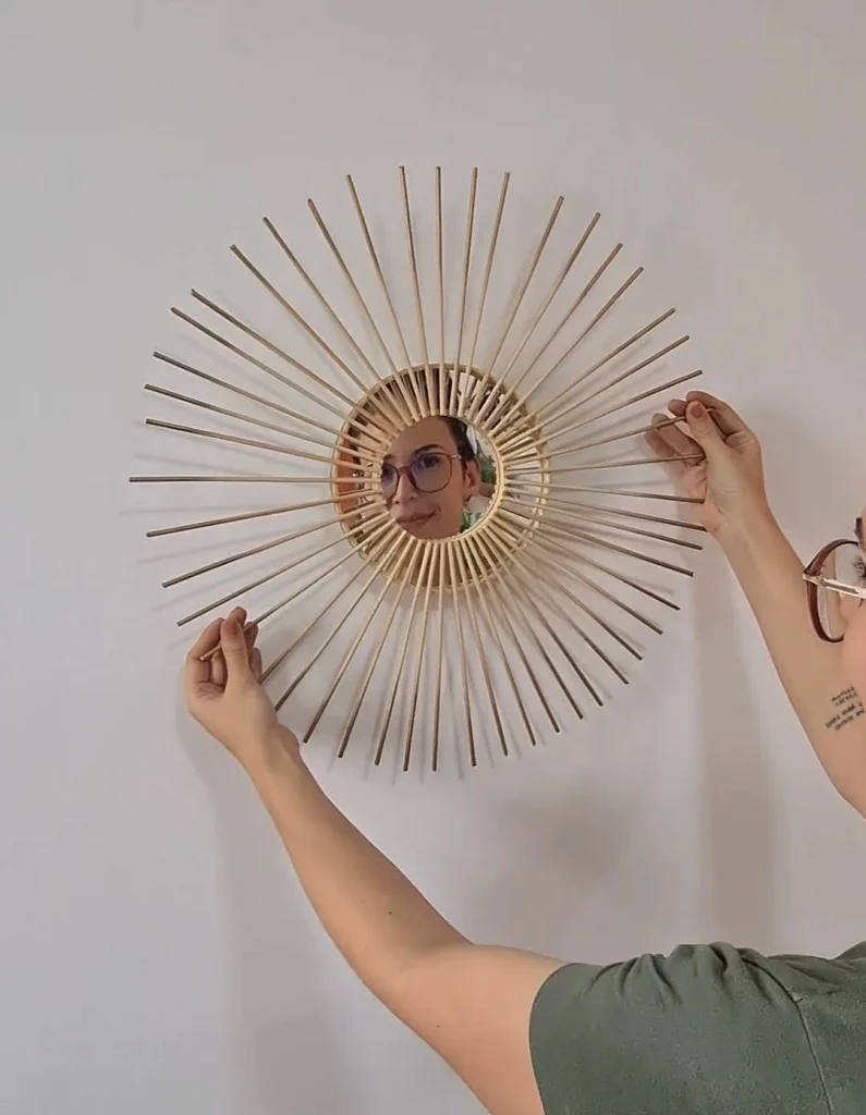 Parede sendo decorada com um espelho redondo que tem raios de sol pintados de dourado, refletindo uma mulher