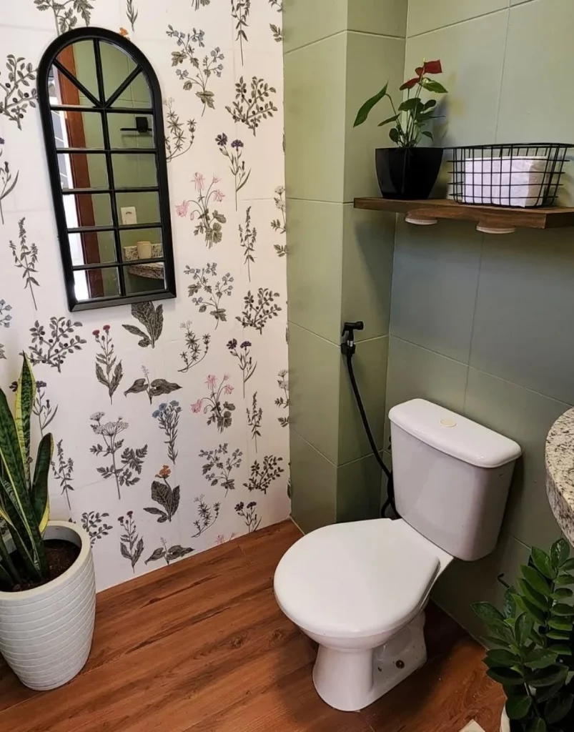 Lavabo decorado com papel de parede floral, prateleira acima do vaso sanitário, espelho em formato de janela e vaso de planta grande.