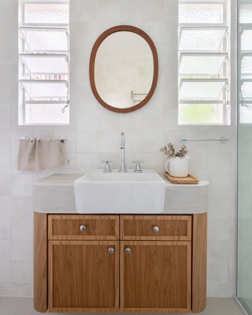 Lavabo minimalista e clean com espelho oval em moldura de maderia, janelas na lateral da pia e armário de madeira.