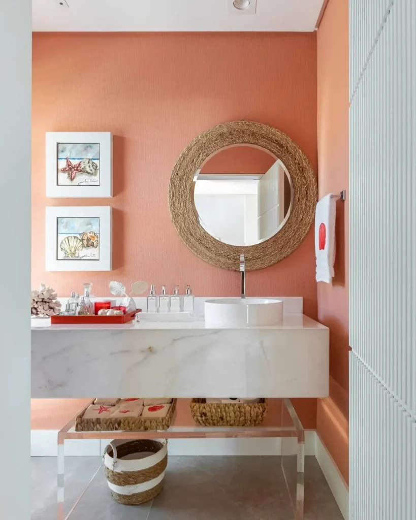 Parede laranja com quadros em aquarela ao lado da pia do lavabo, espelho redondo com decoração de palha e prateleira abaixo da pia.