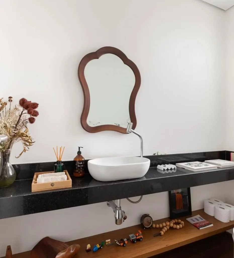 Lavabo com espelho de moldura vintage, bancada de pia comprida e preta com vaso de flores, prateleira abaixo da pia.
