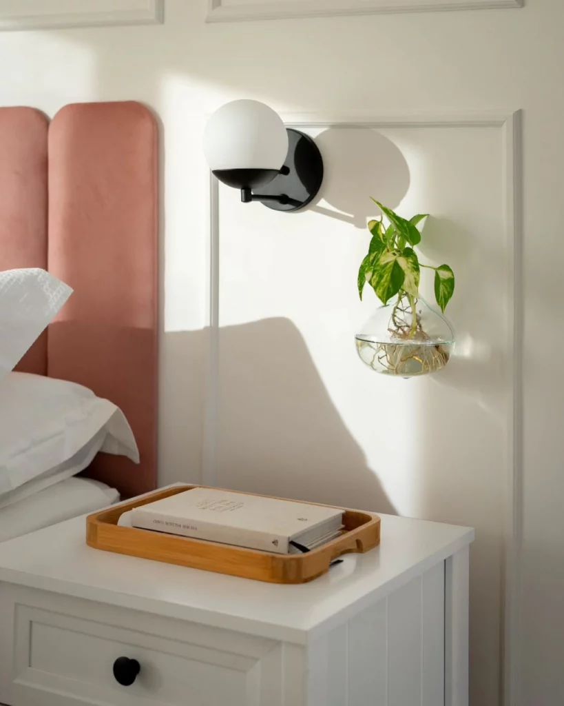 Parede branca de quarto decorada com arandela, boiserie e vaso de planta transparente próximo de uma mesa de cabeceira branca