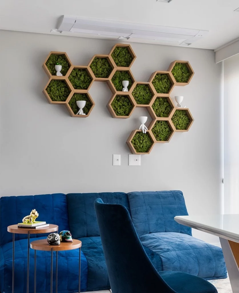 Parede cinza claro decorada com nichos em formato de colmeia, que estão cheios de plantas no fundo. Também tem uma um sofá grande e azul marinho acompanhado de duas mesas pequenas e redondas.