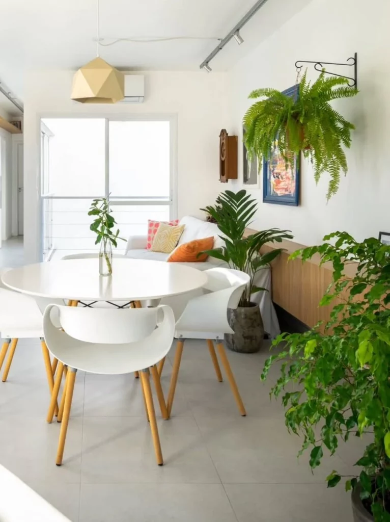 Sala de estar compartilhada com sala de jantar, onde há uma mesa branca central com cadeiras brancas. Muitas plantas no ambiente, incluindo uma samambaia na parede.