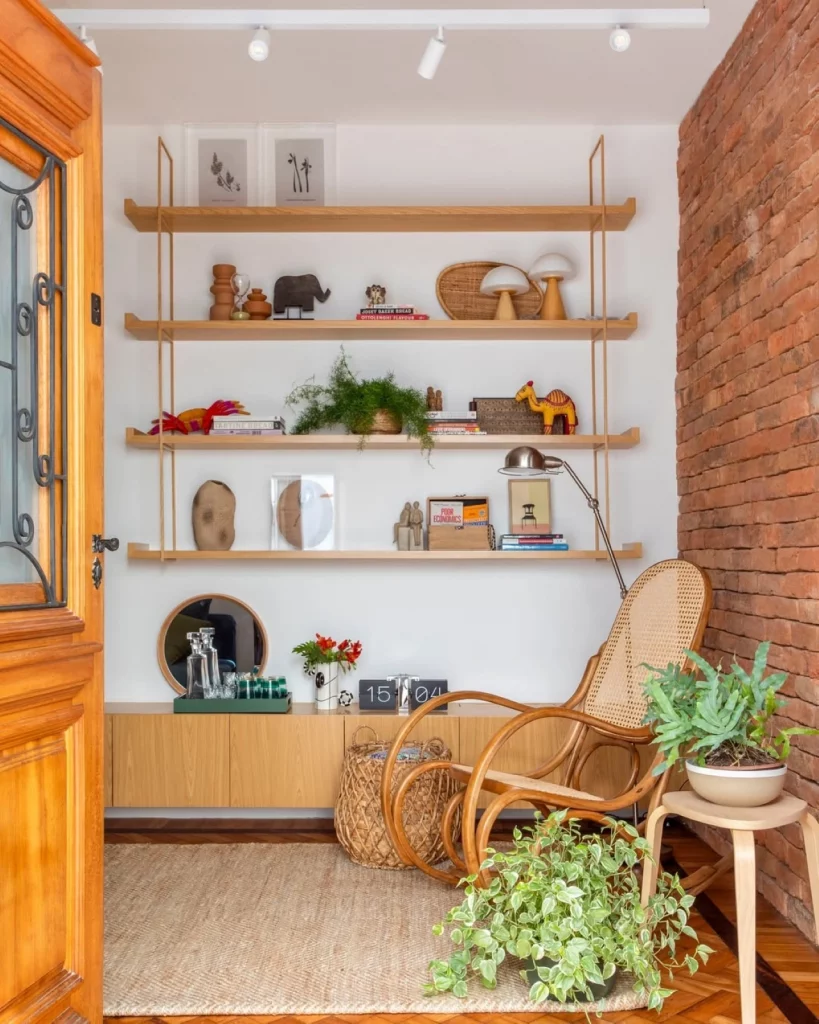 Parede decorada com quatro prateleiras de madeira clara envernizada e muitos objetos decorativos, com plantas e esculturas. Também há uma cadeira de balanço de madeira e uma mesinha ao lado com plantas.
