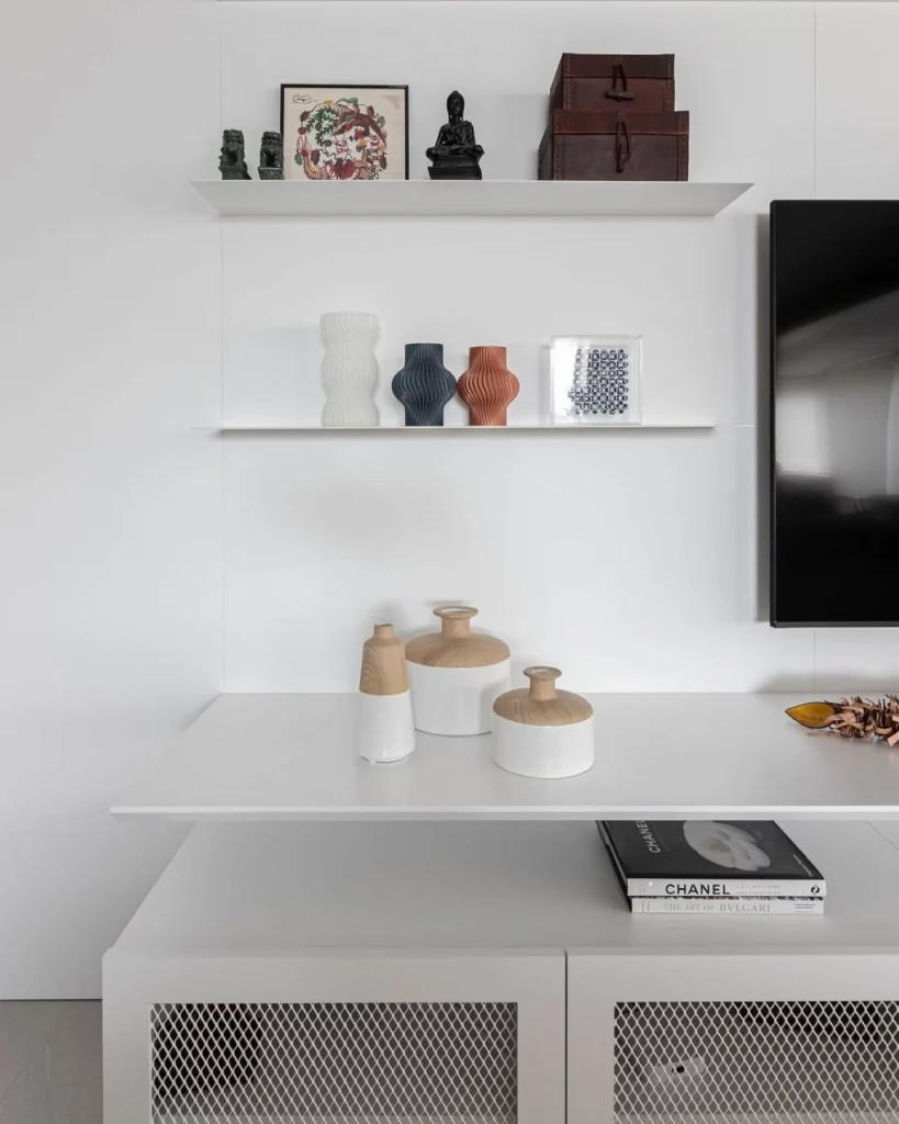 Parede decorada com prateleira branca, onde estão algumas escuclturas, vasos e quadros