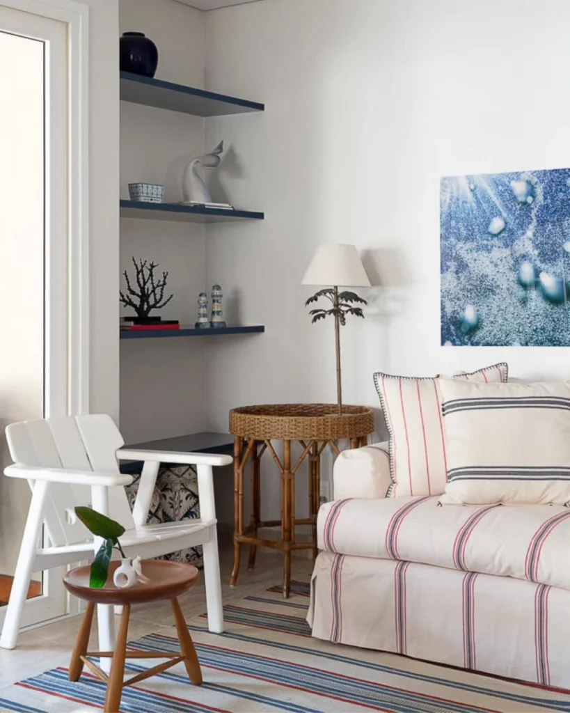 Sala de estar aconchegante, com cadeira branca e sofá listrado, também há um quadro azul na parede, uma mesinha redonda com abajur e três prateleiras azuis marinho no canto com esculturas praianas.