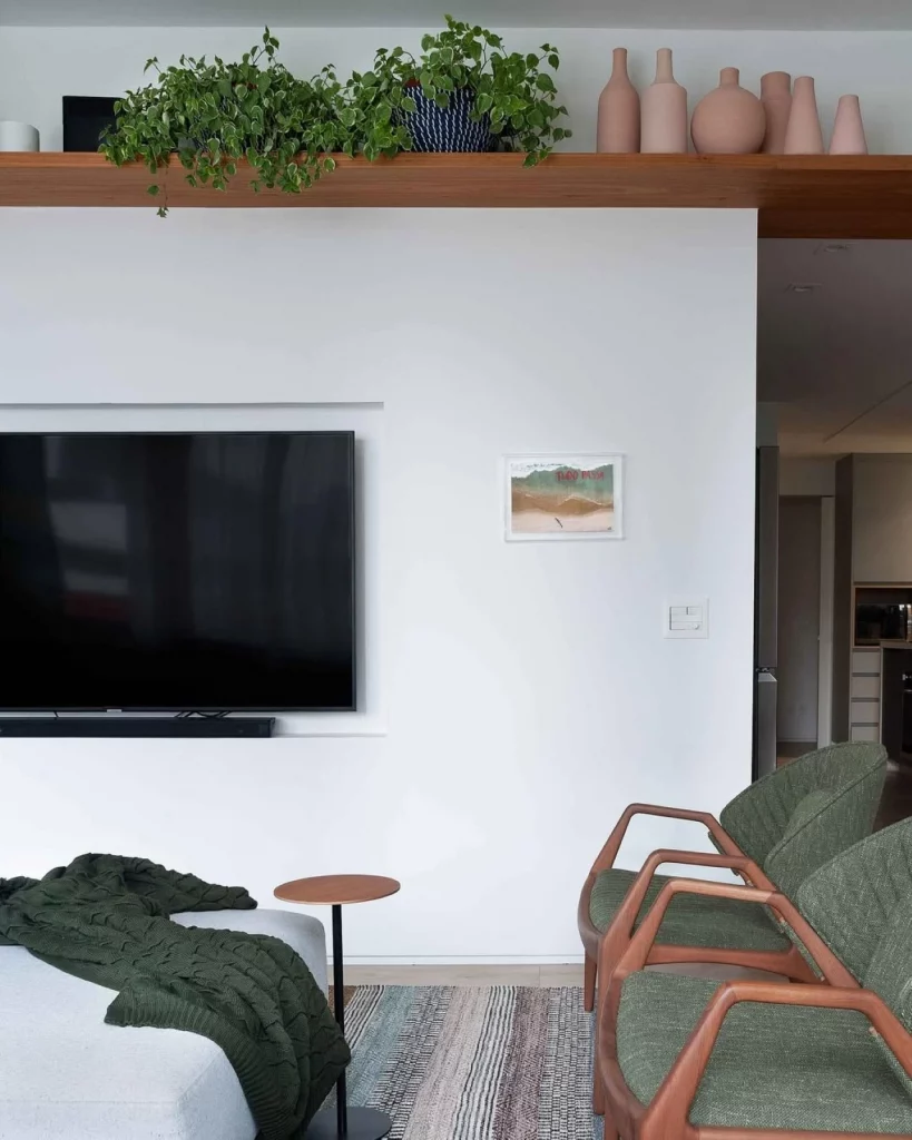 Sala de televisão com um quadro minimalista na parede, poltronas verde musgo e prateleira alta de madeira decorada com plantas e vasos