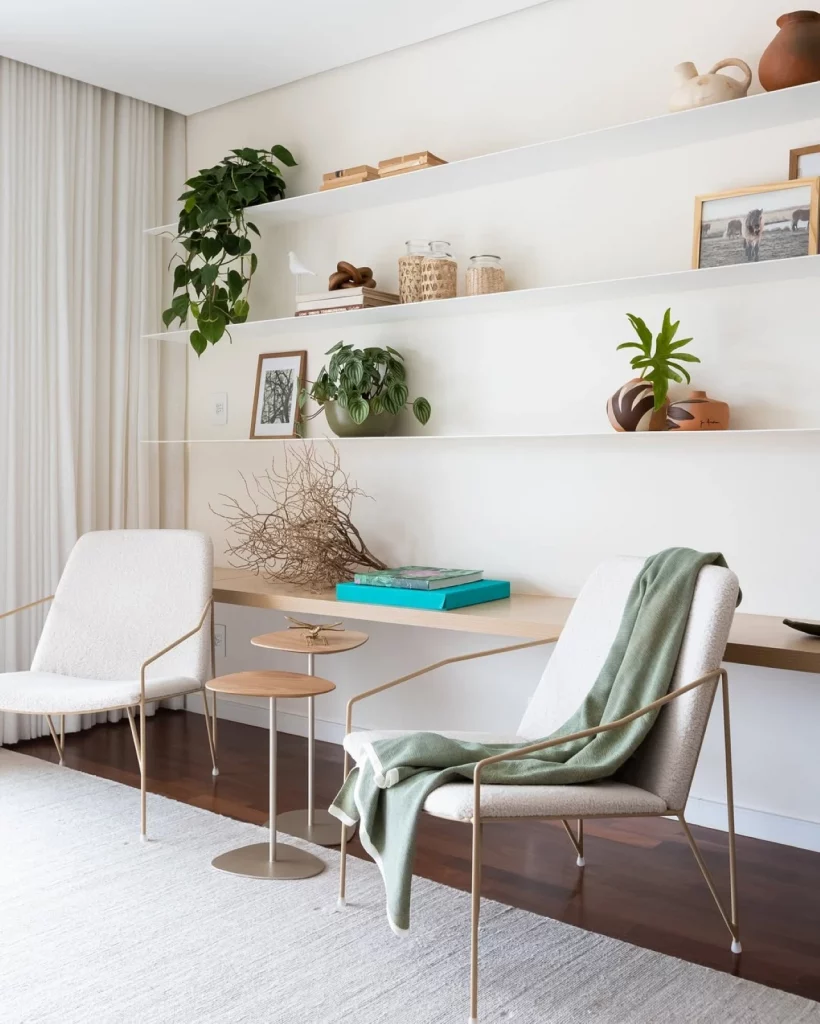 Prateleiras brancas e extensas decoradas com plantas, vasos pequenos e quadros adornando a sala, que tem duas cadeiras brancas e modernas