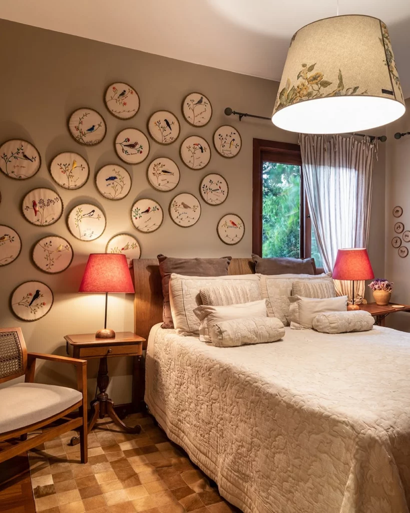 Quarto de casal com bastidores bordados de pássaros ocupando boa parte da parede, também há jogo de luz com abajures nas mesas de cabeceira e uma cama com colcha clara.