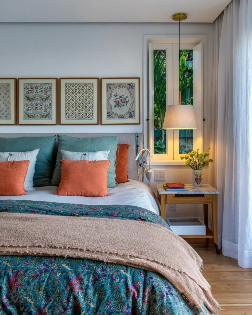 Quarto de casal aconchegante com jogo de cama colorido e almofadas de tamanhos diferentes. Na parede há quadros enfileirados em linha reta e com diferentes desenhos.