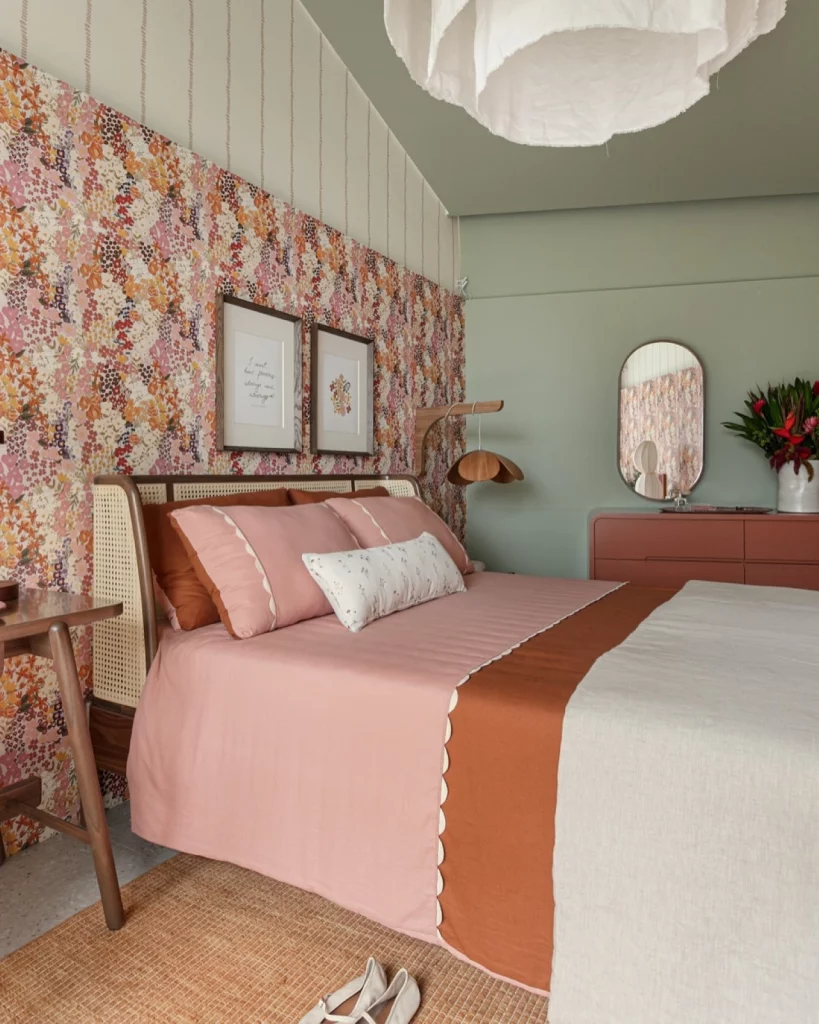 Quarto de casal aconchegante e delicado, com papel de parede floral em tons de rosa, quadros na parede e roupa de cama em tons de rosa, terracota e off white.