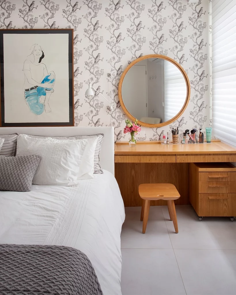 Quarto com papel de parede floral em tons sóbrios e estilo vintage, também há um quadro na parede e um espelho redondo na penteadeira.