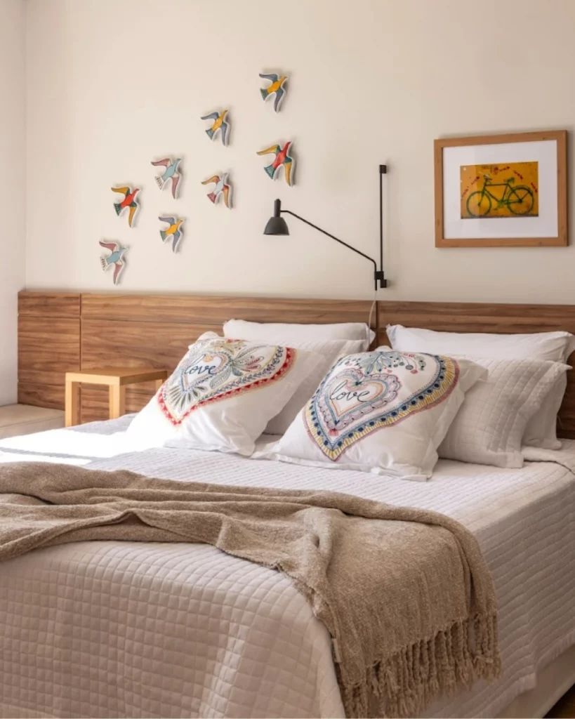 Quarto de casal confortável com escultura de pássaros coloridos voando na parede, quadro e arandela na parede. Na cama, há almofadas estampadas e uma manta.