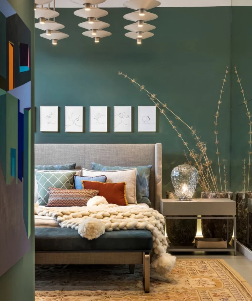 Quarto de casal sofisticado, com parede pintada em tom de verde escuro e frio, tem quadros próximos da cabeceira, que estão enfileirados em linha reta. Também há uma manta fofa na cama e um vaso de com bambu próximo da mesa de cabeceira.