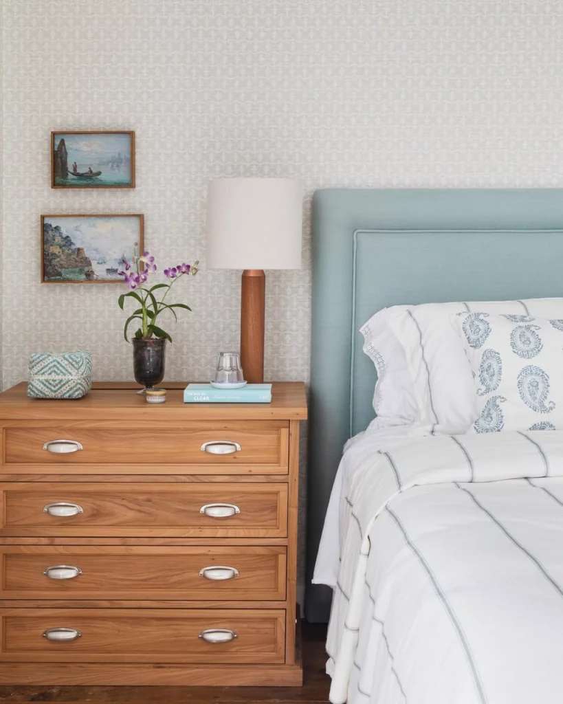 Quarto aconchegante e delicado, com papel de parede em tons claros de bege e estampa suave; na parede també há quadros e uma cabeceira azul claro apoiada. Há uma mesa de cabeceira de madeira, com abajur e vaso de planta.