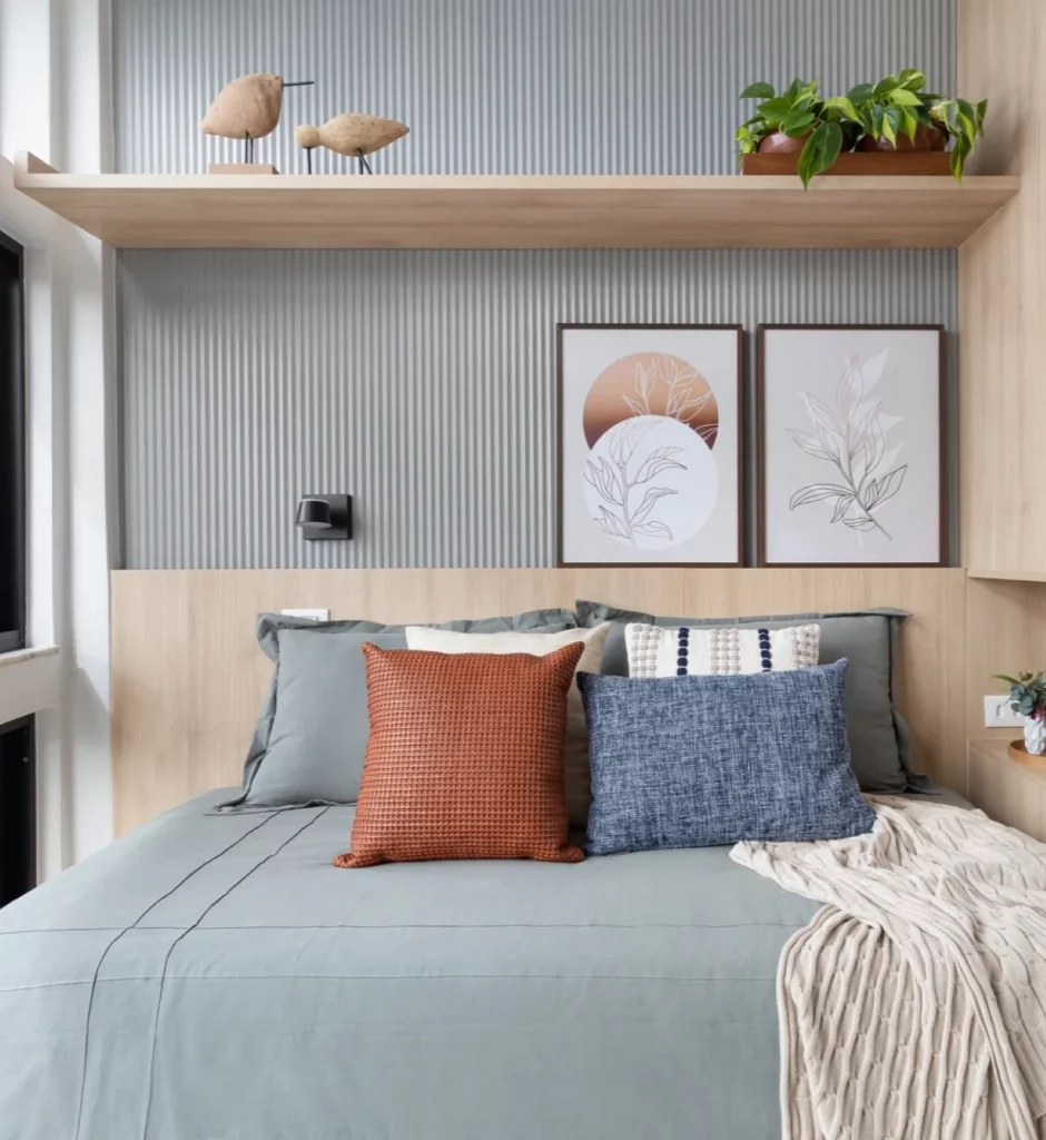Quarto de casal pequeno, com cama grande, parede com prateleira com esculturas e vaso de planta, quadros apoiados na cabeceira e jogo de cama colorido com almofadas de tamanhos diferentes e cores sérias.