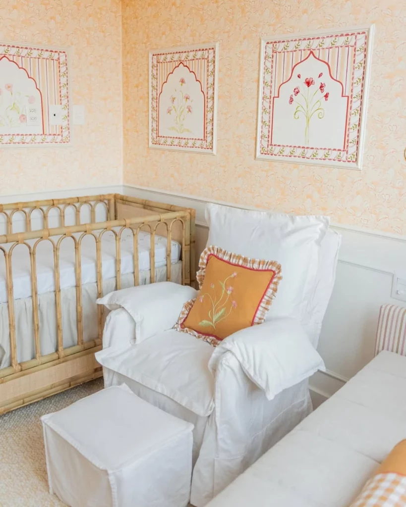 Quarto de bebê feminino, com quadros de flores delicadas na parede e papel de parede amarelo, parede com boiserie branco e poltrona de amamentação branca com almofada laranja e flor bordada.