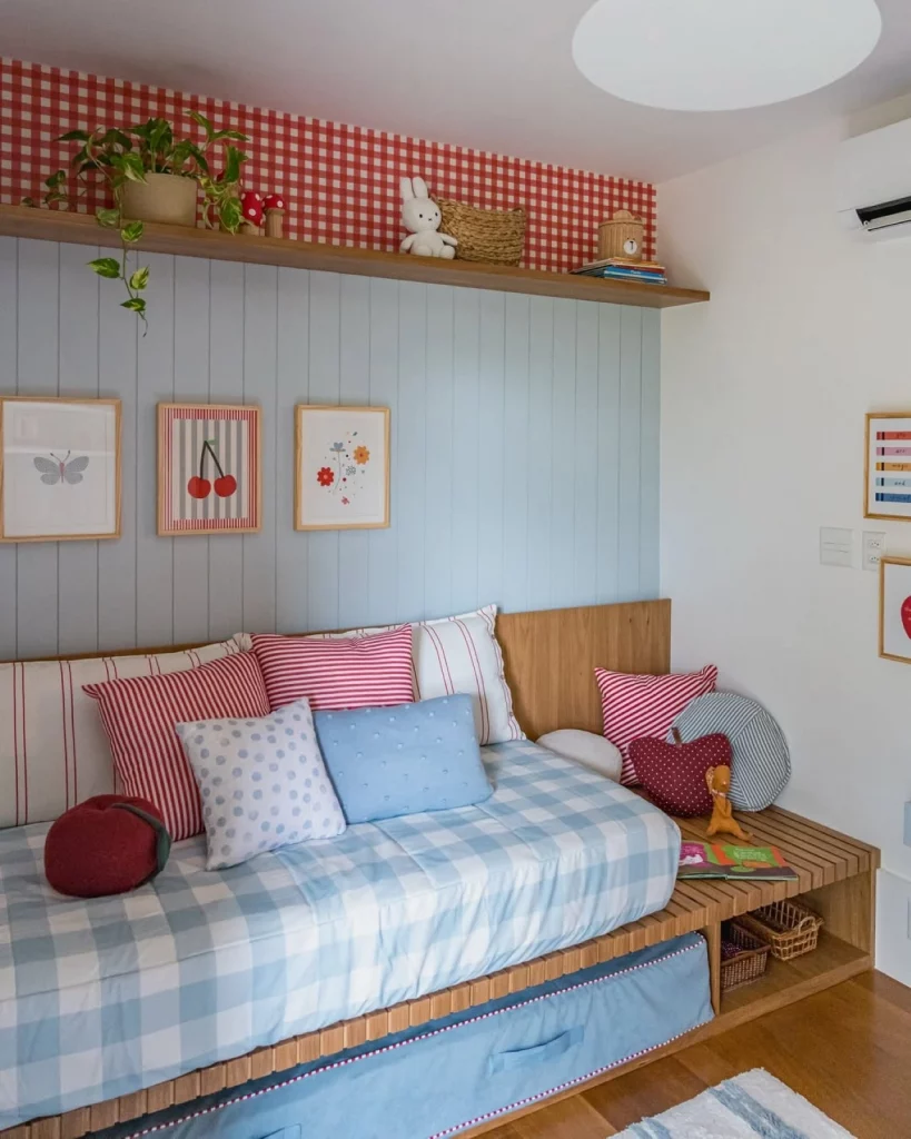 Quarto infantil de menina, com parede ripada azul, papel de parede xadrez vermelho, trio de quadros, almofadas coloridas na cama, prateleira com planta.