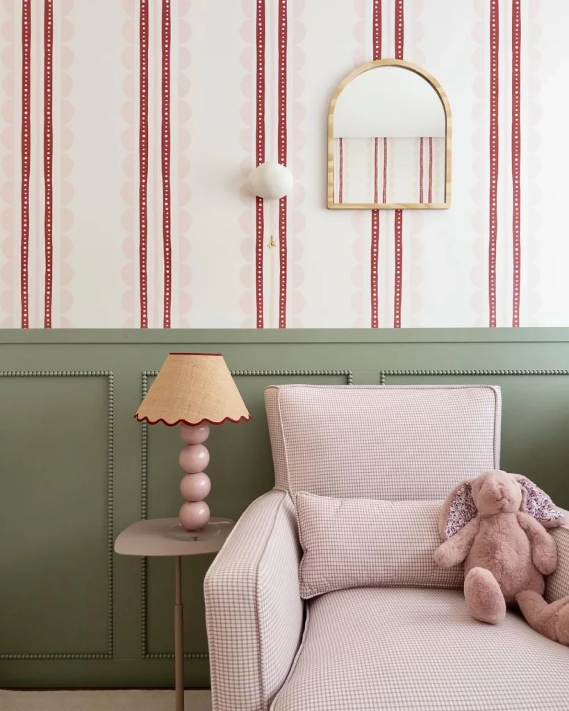Quarto infantil feminino com boiserie verde na parede, papel de parede listrado veremhlo, espelho em arco pequeno, poltrona rosa e abajur delicado.