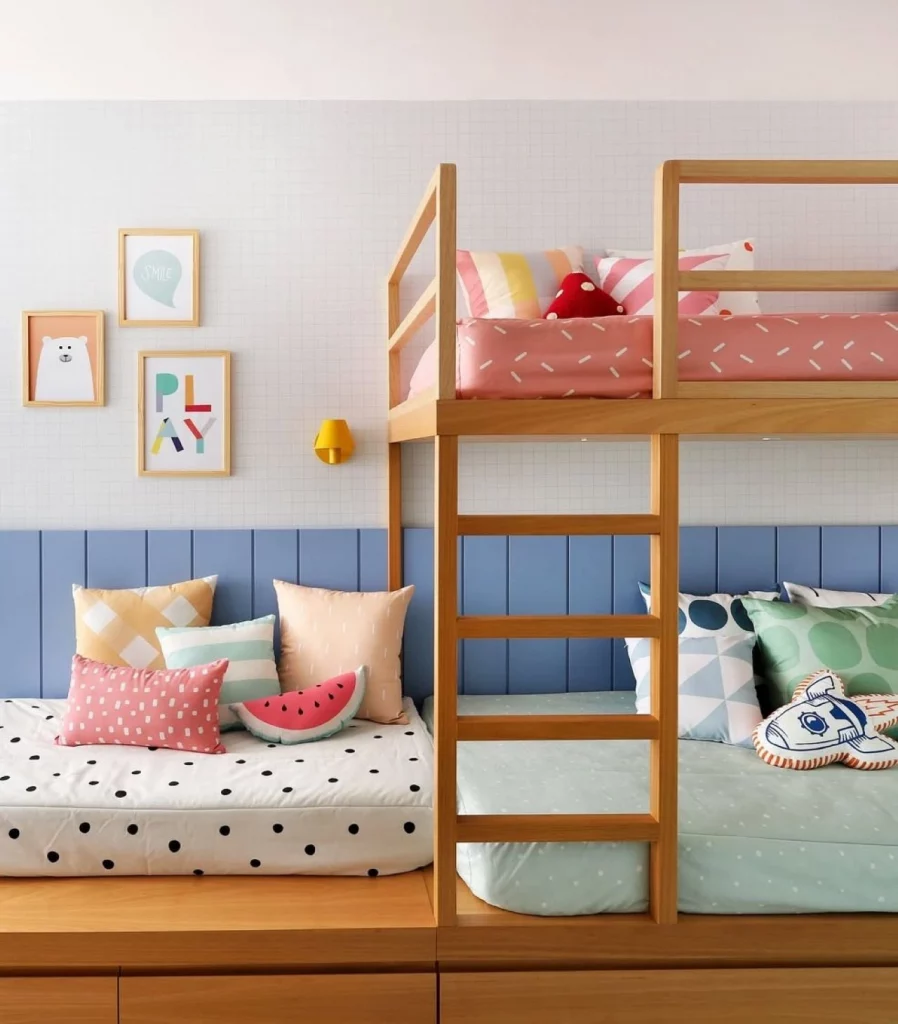 Quarto infantil de menina colorido, com beliche, ripado azul em metade da parede, com pintura segmentada azul claro e trio de quadros; na cama tem almofadas divertidas e de diversos formatos.