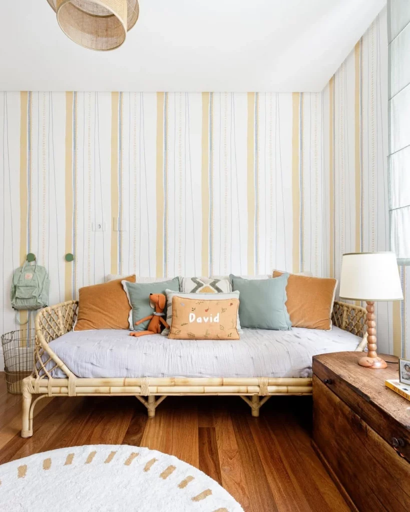 Quarto infantil de menino, com papel de parede listrado e amarelo pastel, cama baixa com muitas almofadas e mesa de cabeceira de madeira com abajur.