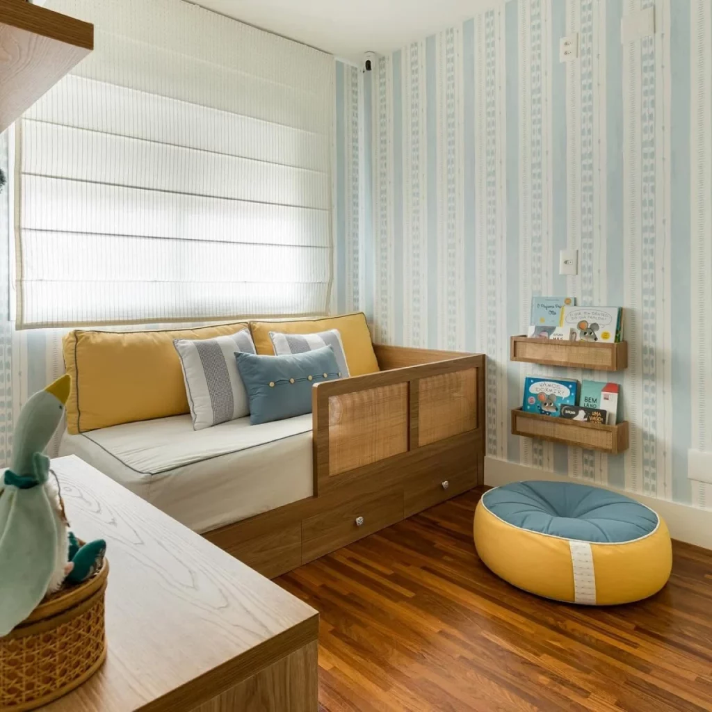 Quarto infantil de menino com papel de parede listrado azul, cama com grade de madeira e palha indiana, puff amarelo com azul e prateleiras de livros baixas de madeira na parede.