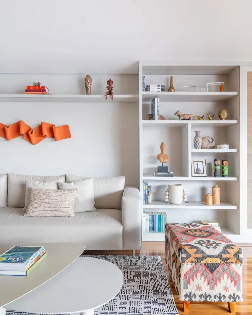 Sala de estar decorada com prateleira e estante branca, também há um sofá branco e um puff retangular estampado; o destaque da decoração é uma escultura laranja na parede.