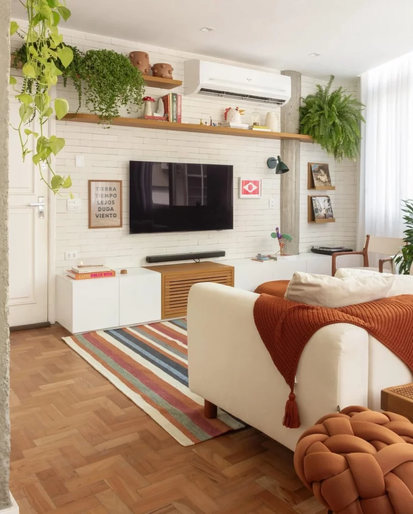 Sala de televisão decorada com prateleiras, onde estão muitas plantas e objetos decorativos. Há um sofá branco com uma manta avermelhada e escura.
