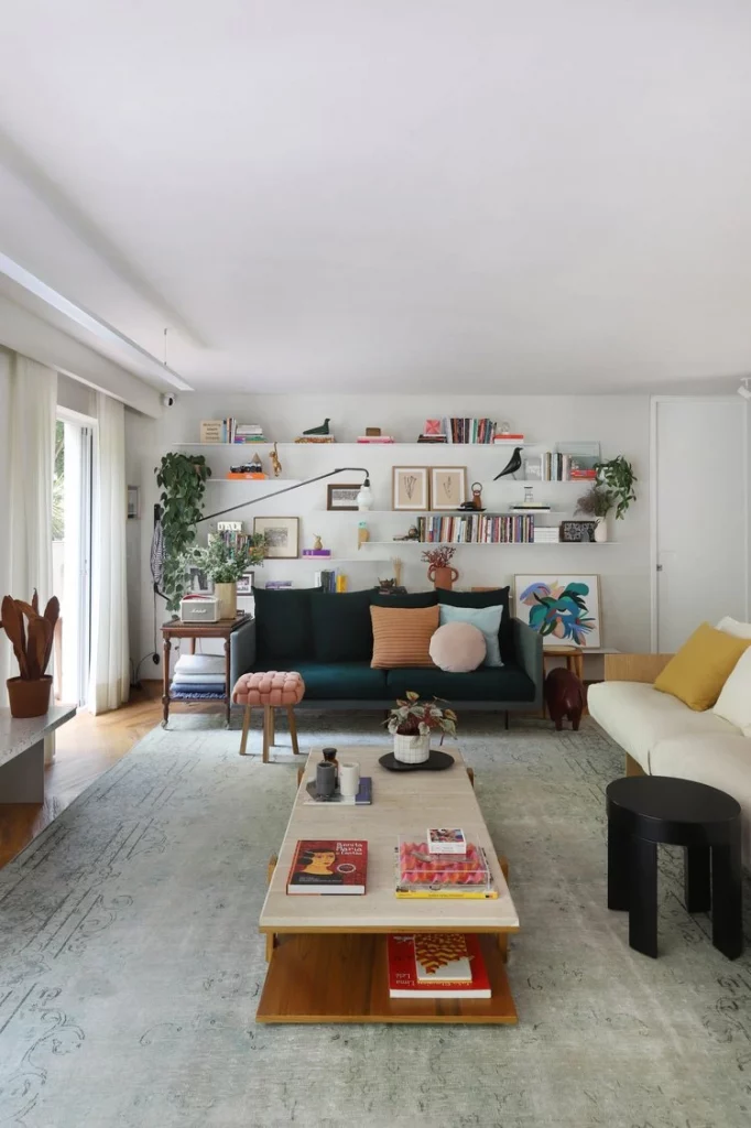 Sala de estar aconchegante e grande, com parede decorada com prateleiras e acessórios como livros, quadros, plantas e esculturas; também há uma mesa de centro baixa e um sofá azul marinho com almofadas coloridas.