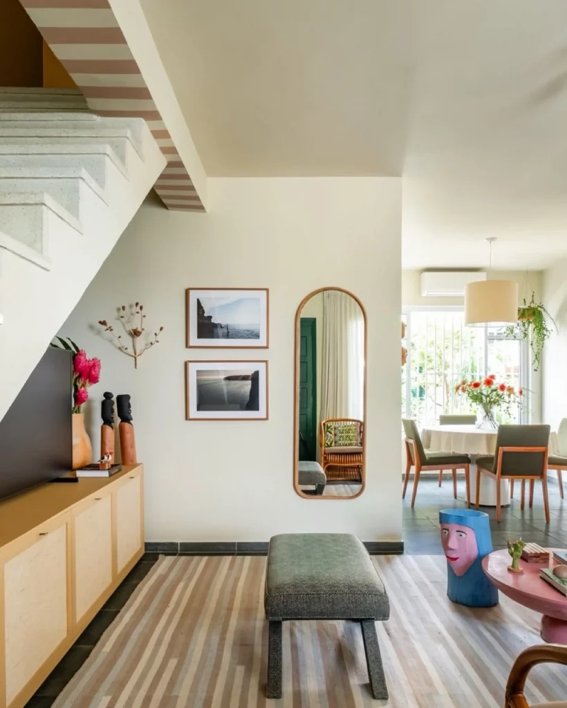 Cantinho da parede da sala decorado com espelho em formato de arco e quadros, também há um banco acolchoado no centro com uma escultura próxima.