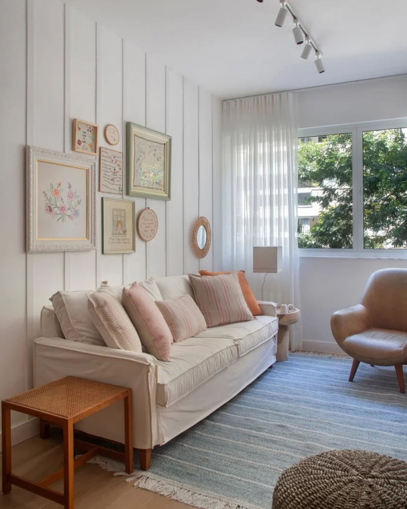 Sala de estar clean, com parede com ripas de madeira brancas e quadros de diferentes tamanhos e formatos; também há um sofá branco com almofadas e uma poltrona próxima.