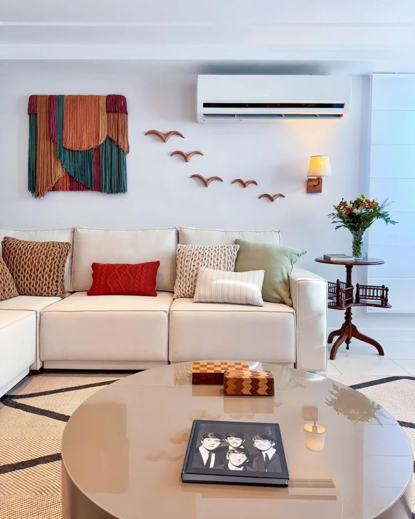 Sala de estar aconchegante e vibrante, com esculturas de pássaros voando na parede e quadro colorido texturizado de tecido. Também há um sofa branco com almofadas coloridas de tamanhos diferentes, mesa central e mesa lateral.