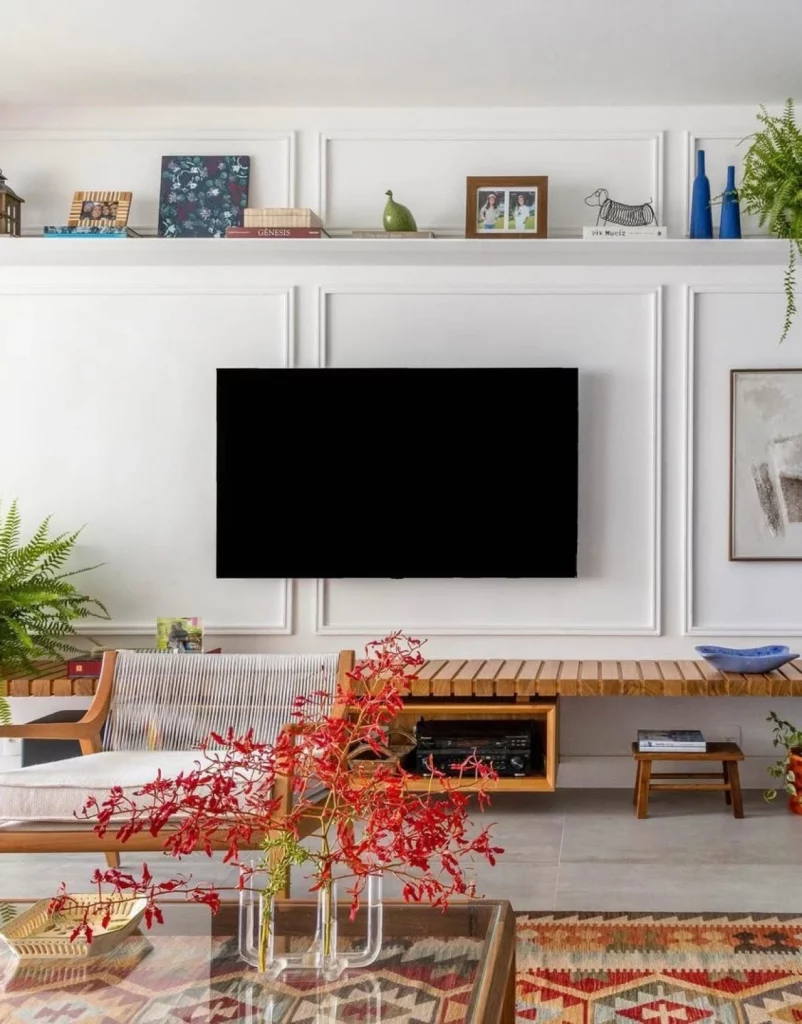 Sala de estar com boiserie, prateleira com plantas, quadros, porta-retratos e esculturas. Há também uma poltrona baixa e um vaso com planta vermelha chamativa.