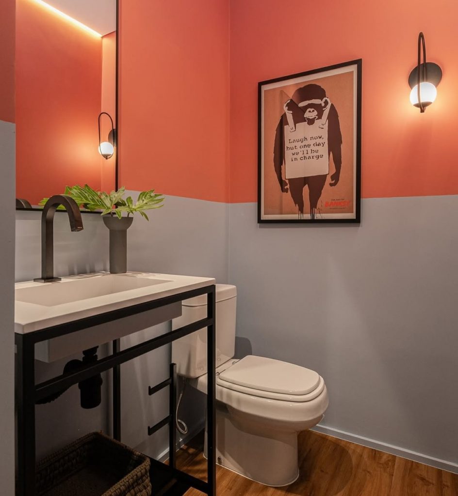 Banheiro com estética divertida e leve, com quadro de macaco pendurado, paredes cinza com laranja e arandela.